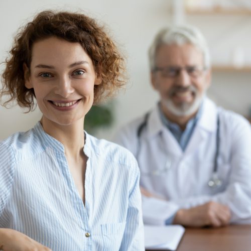 Happy healthy young woman patient visiting old male doctor physician sitting at medical appointment consultation in hospital looking at camera. Women healthcare medical checkup concept. Portrait