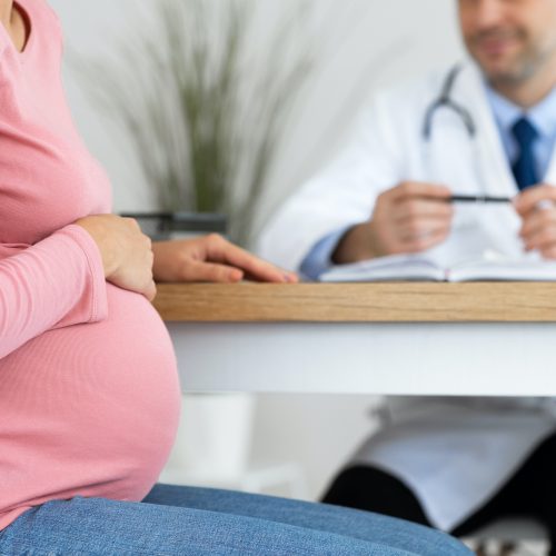 Prenatal Care Concept. Unrecognizable pregnant woman sitting at clinic or hospital, having consulation with male gynecologist, closeup on her hand holding and embracing belly. Visiting obstetrician